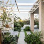 Concrete pavers with grass strips lead through a lush garden toward a white outdoor patio seating area.