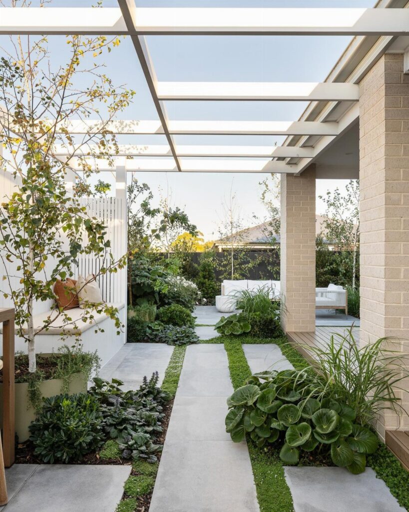 Concrete pavers with grass strips lead through a lush garden toward a white outdoor patio seating area.