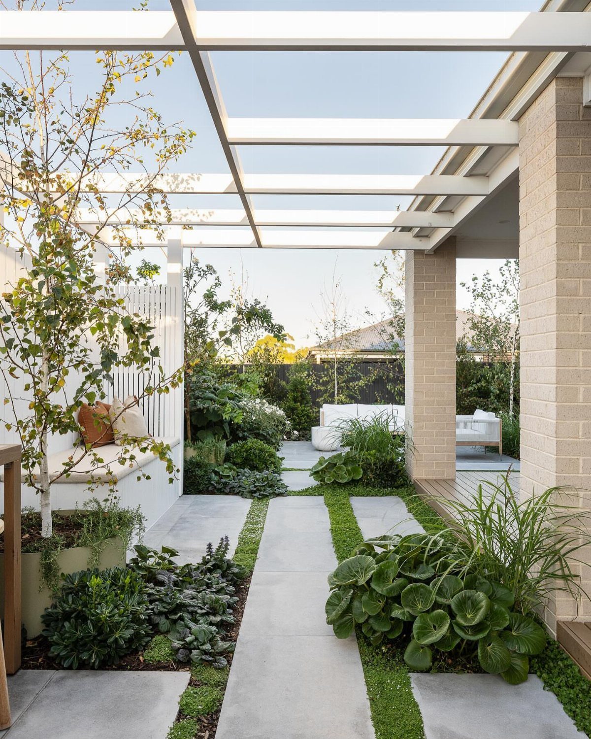Concrete pavers with grass strips lead through a lush garden toward a white outdoor patio seating area.