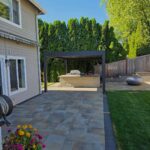 Stone patio pavers leading toward a dark pergola and an outdoor kitchen area.