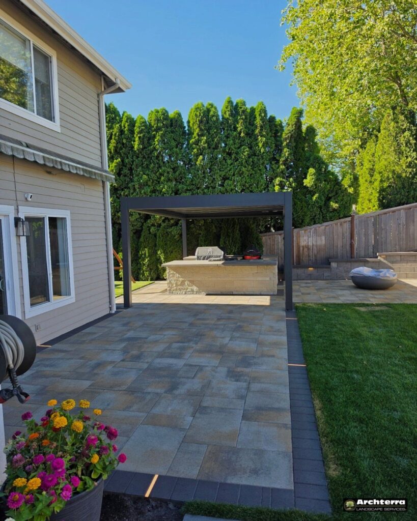 Stone patio pavers leading toward a dark pergola and an outdoor kitchen area.