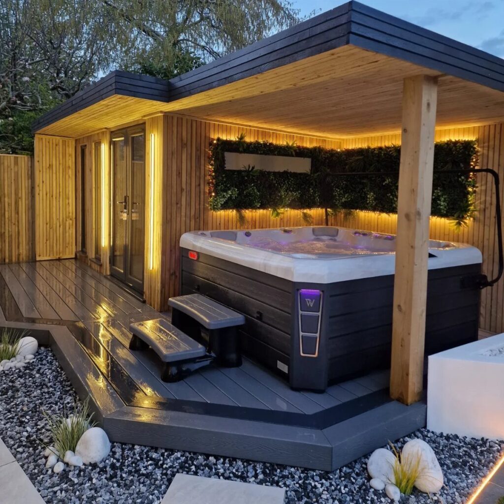 A modern wooden gazebo housing a lit hot tub on a dark grey deck with greenery.