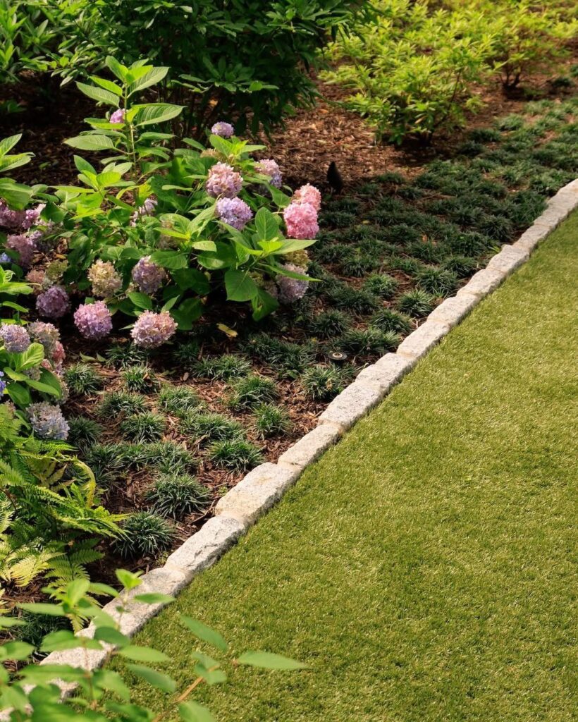 Stone edging bordering a lush green lawn and a garden bed with pink hydrangeas.
