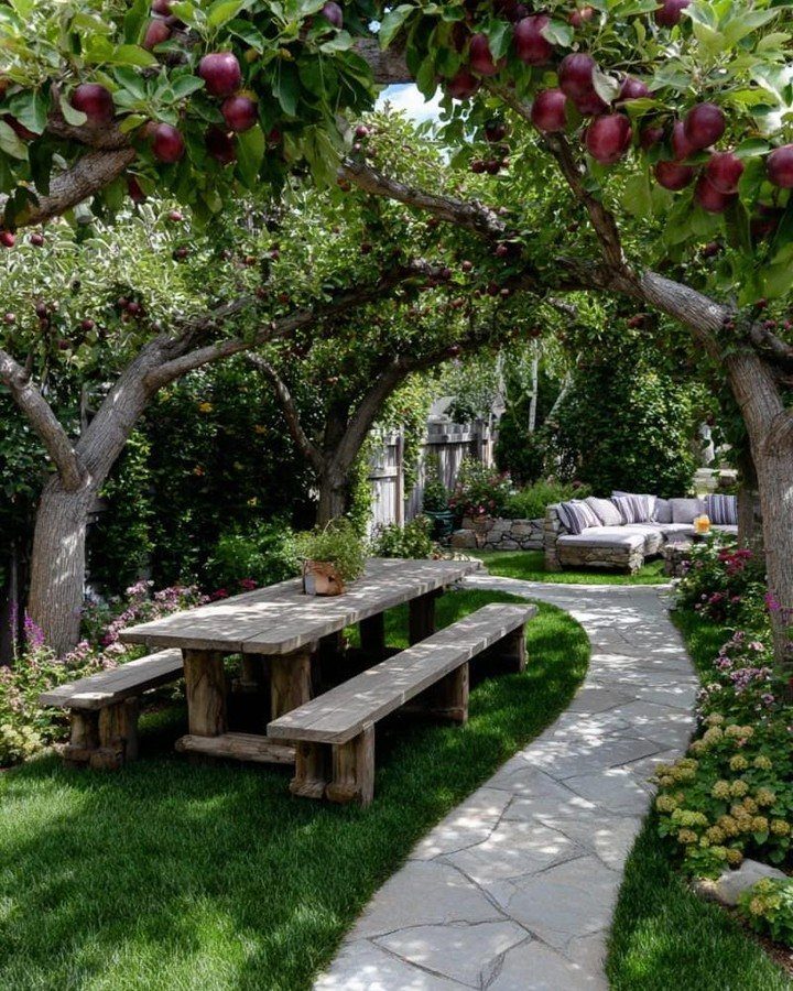A wooden dining table and benches sit under apple trees beside a stone garden path.