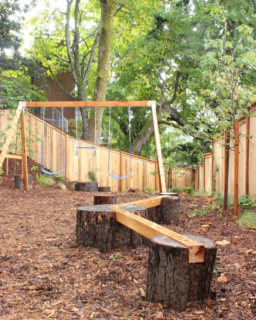 Wooden swing set and tree stump balance beam in a mulched backyard play area.