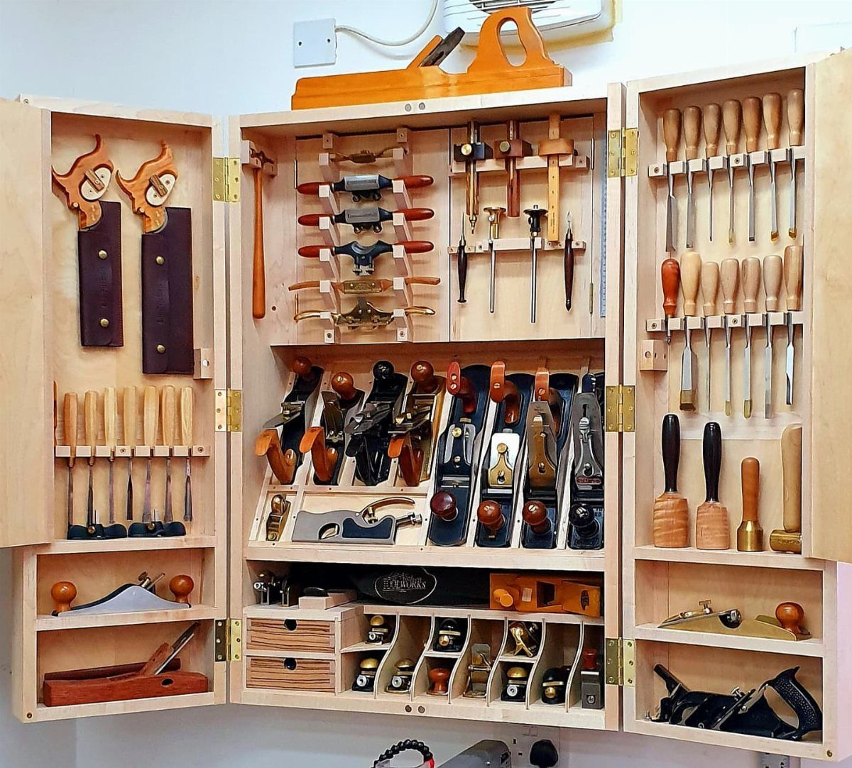 An open wooden wall cabinet organized with various woodworking hand tools and compartments.