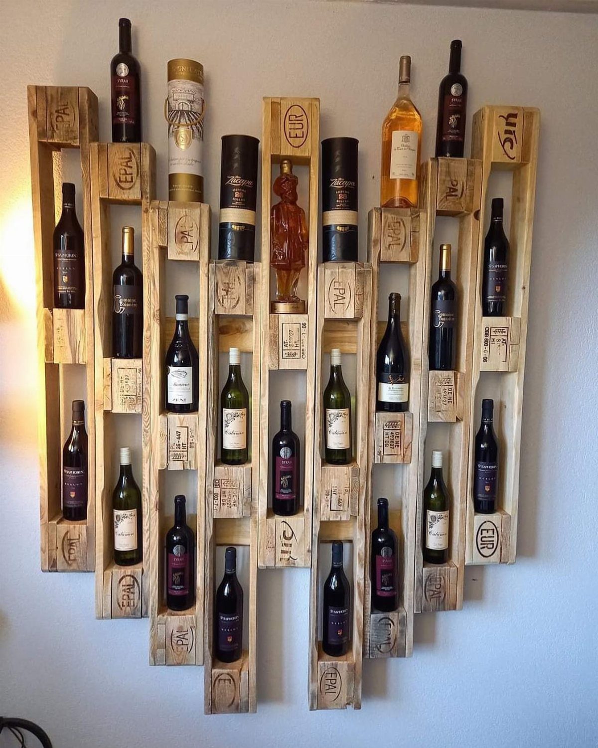 A wooden pallet wine rack mounted on a white wall holding many wine bottles.